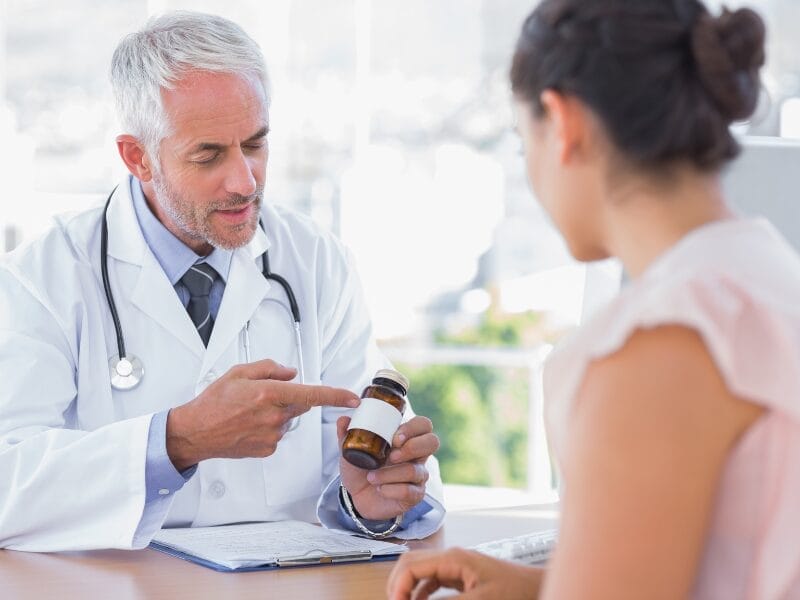 A doctor in a white coat shows a medication bottle to a patient seated across a desk in a medical office.